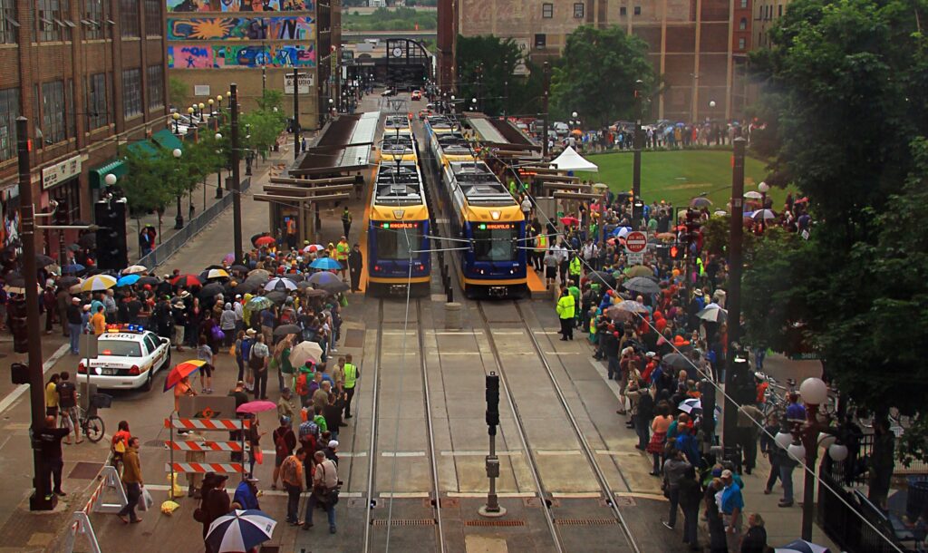 Image of the Opening Day of the Green Line in Saint Paul, MN. Two light rail trains are situated side by side and surrounded by a larger celebratory crowd of people. 