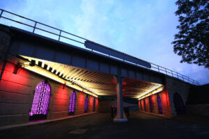 Image of over.under.pass in Greensboro, NC. The underpass is lit with red, purple, and white lights creating a friendly environment for people walking, rolling, or using the space.