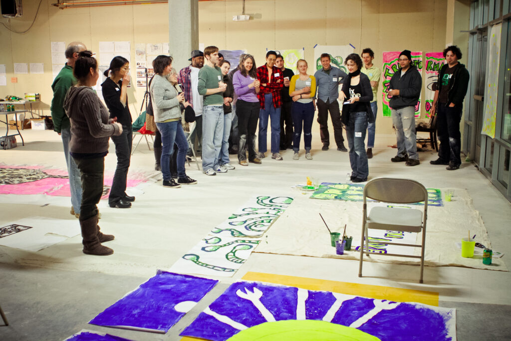 Image of a group of people standing in a semi-circle inside a building around two large pained banners on the ground.