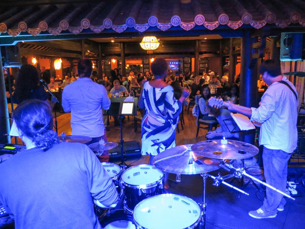 Image of a musical group performing inside a building. Photo is taken from behind and there is a blue lighting covering the performers.