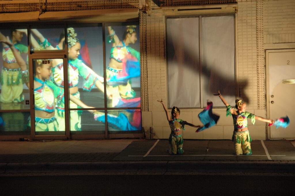 Image of outside dance performance. On the left is a colorful projection of dancers against the wall of a building. On the right are two dancers with their arms raised.