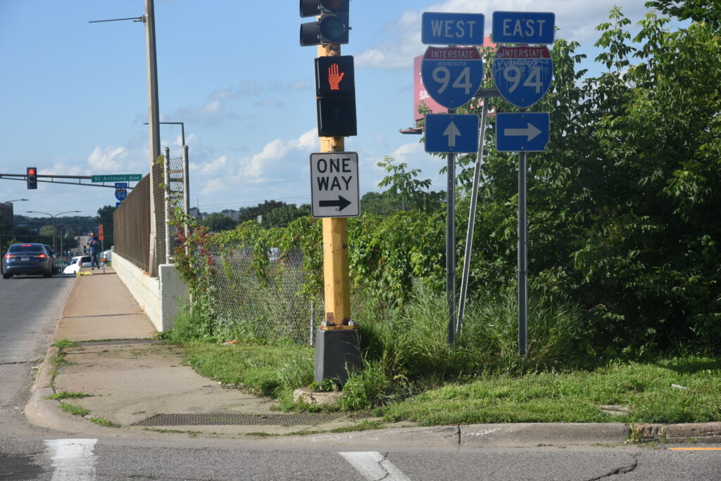 Image of the corner of a bridge over I-94 in the Rondo neighborhood of Saint Paul, MN. There are two signs, one for I-94 West and one for I-94 East.