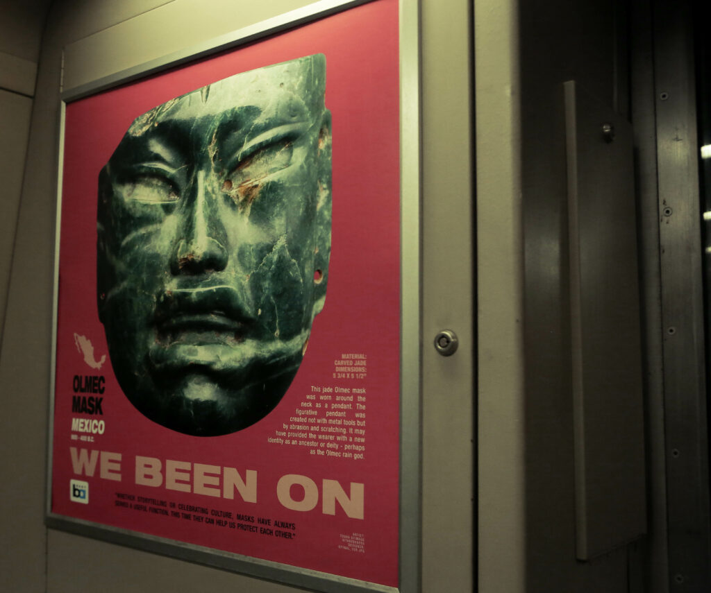 Image of a pink poster on a train car that reads “We been on” and features a slate colored cultural mask.
