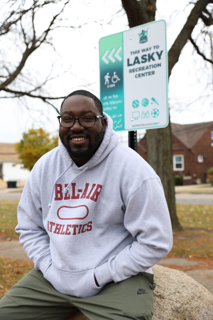 Artist Ndubisi Okoye sits on a boulder in front of a teal and white wayfinding sign featuring the Detroit logo that directs people to nearby Lasky Recreation Center.