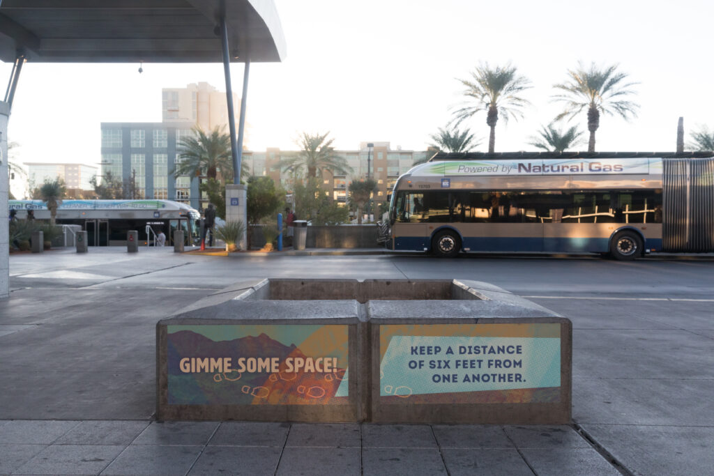 Image of two vinyl signs adhered to a concrete seating area which read “Gimme some space!” and “Keep a distance of 6 feet from one another.” In the background are buses and palm trees.