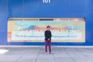 Image of a person standing in front of a sign on an exterior wall of Bonneville Transit Center which reads "A wave is as good as a smile."