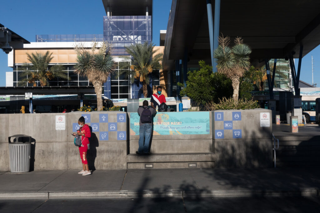 Image of people gathering at a seating area outside of Bonneville Transit Center. Vinyl signage in the seating area includes small blue icons with symbols of buses, people, and footprints and a larger sign that reads “Dude, where is your mask?” and below in smaller font “Masks keep you and others safe.”