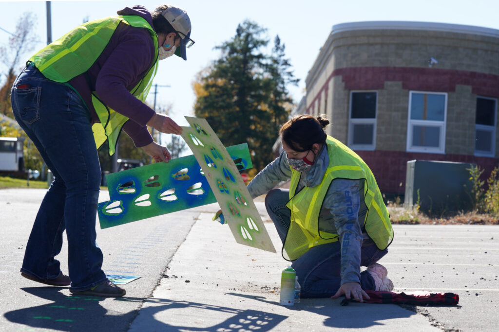 Artist and volunteer use stencils to spray paint green animal footprints on a street.