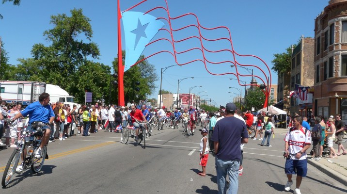 A parade passes under a statue of a Puerto Rican flag in the Paseo Boricua neighborhood of Chicago. The city gifted a matching pair of flag statues to the Puerto Rican neighborhood in 1995. Flickr photo by Emily. https://flickr.com/photos/ebarney/2578770769