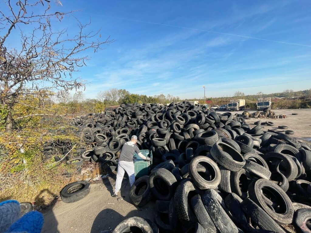 A pile of several illegally dumped tires in a parking lot, pictured against trees in the background and a blue afternoon sky. Photo from Binghampton Development Corporation.